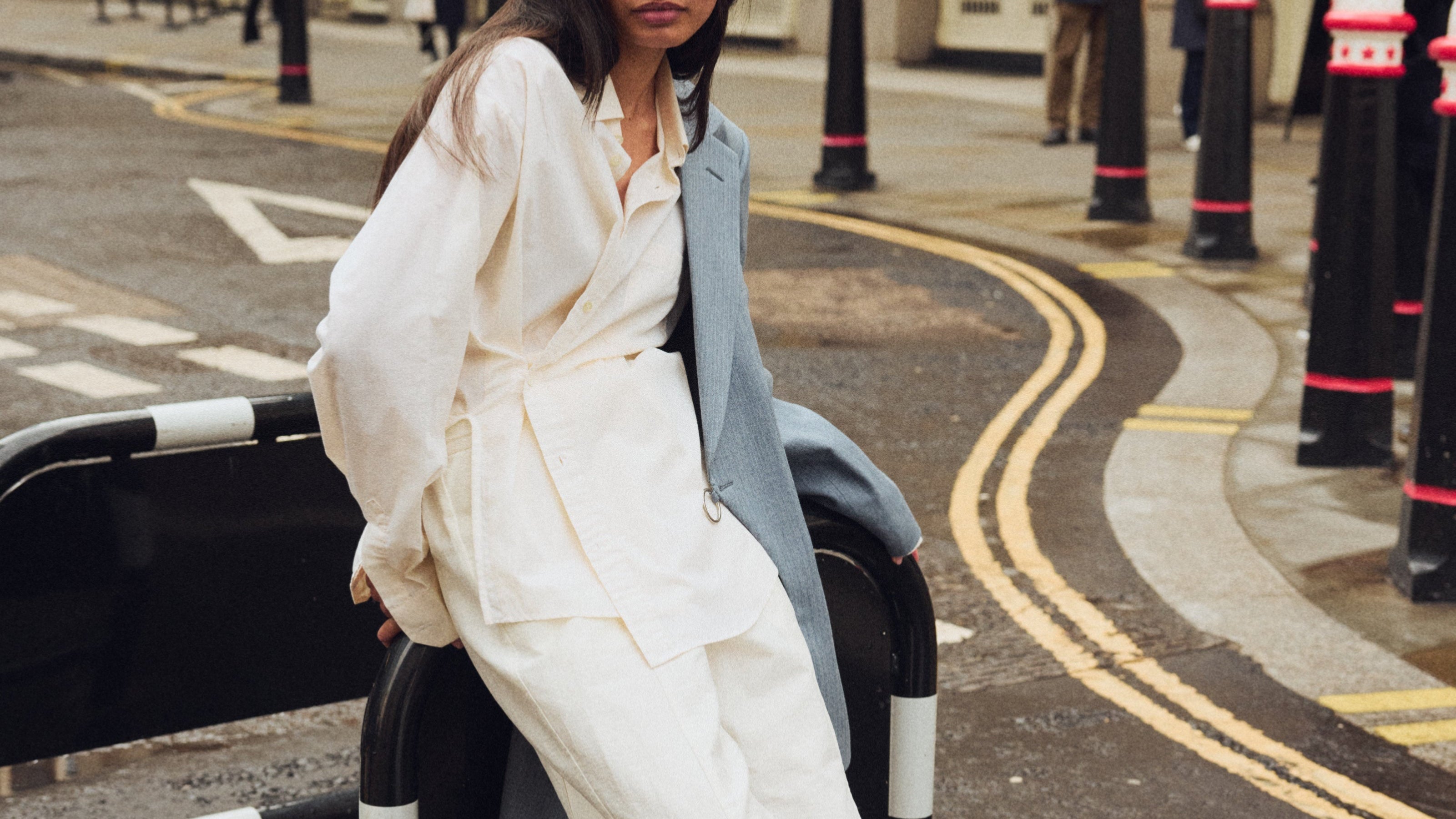 Woman in a white outfit leaning against a black road barrier on a city street.
